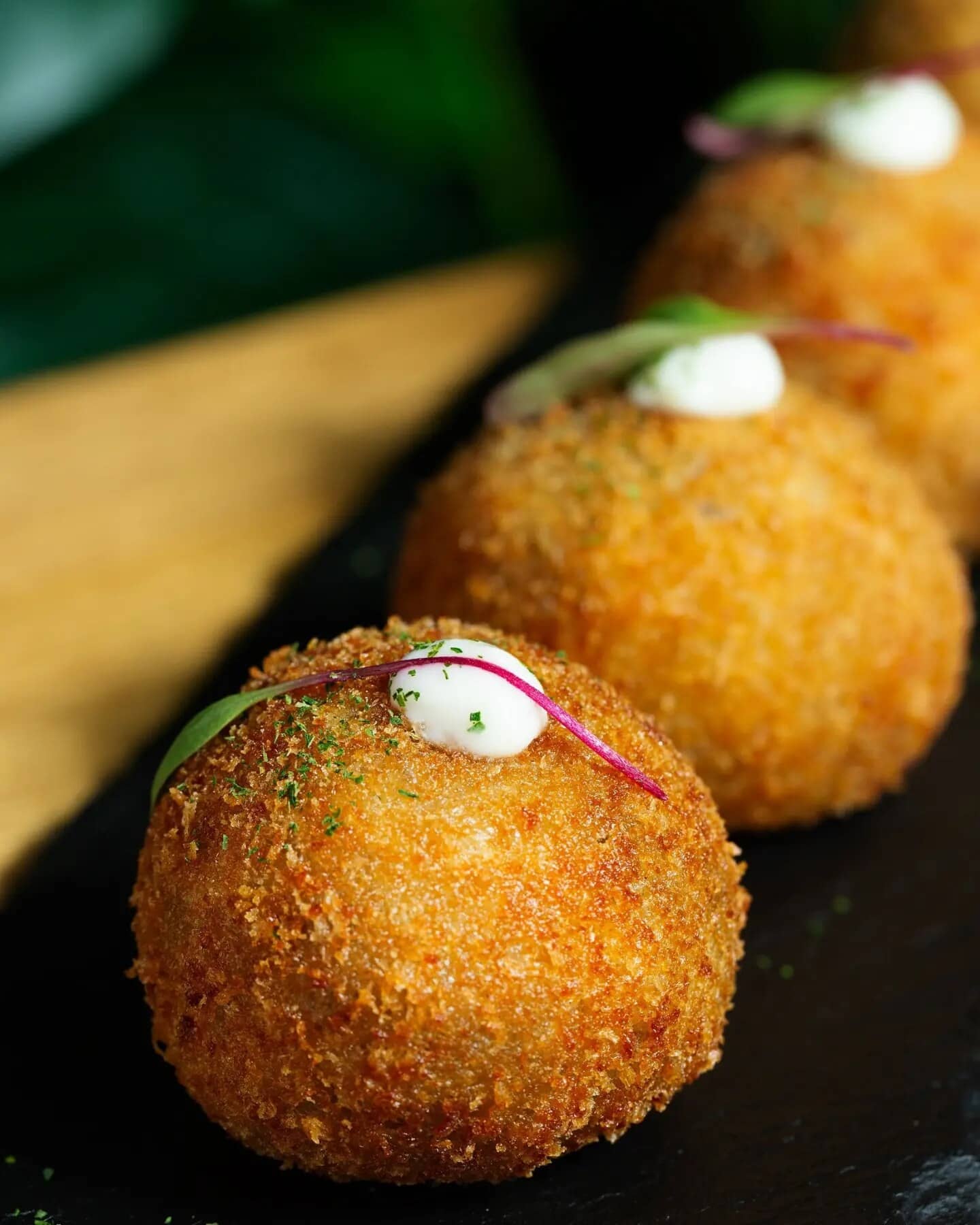 Três arancini de costela empanados, com um ponto de maionese e broto roxo sobre cada um.
