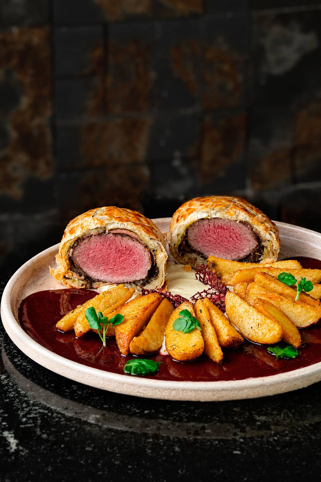 Beef Wellington cut in half, pink tenderloin inside the puff pastry, with rustic potatoes and dark sauce.