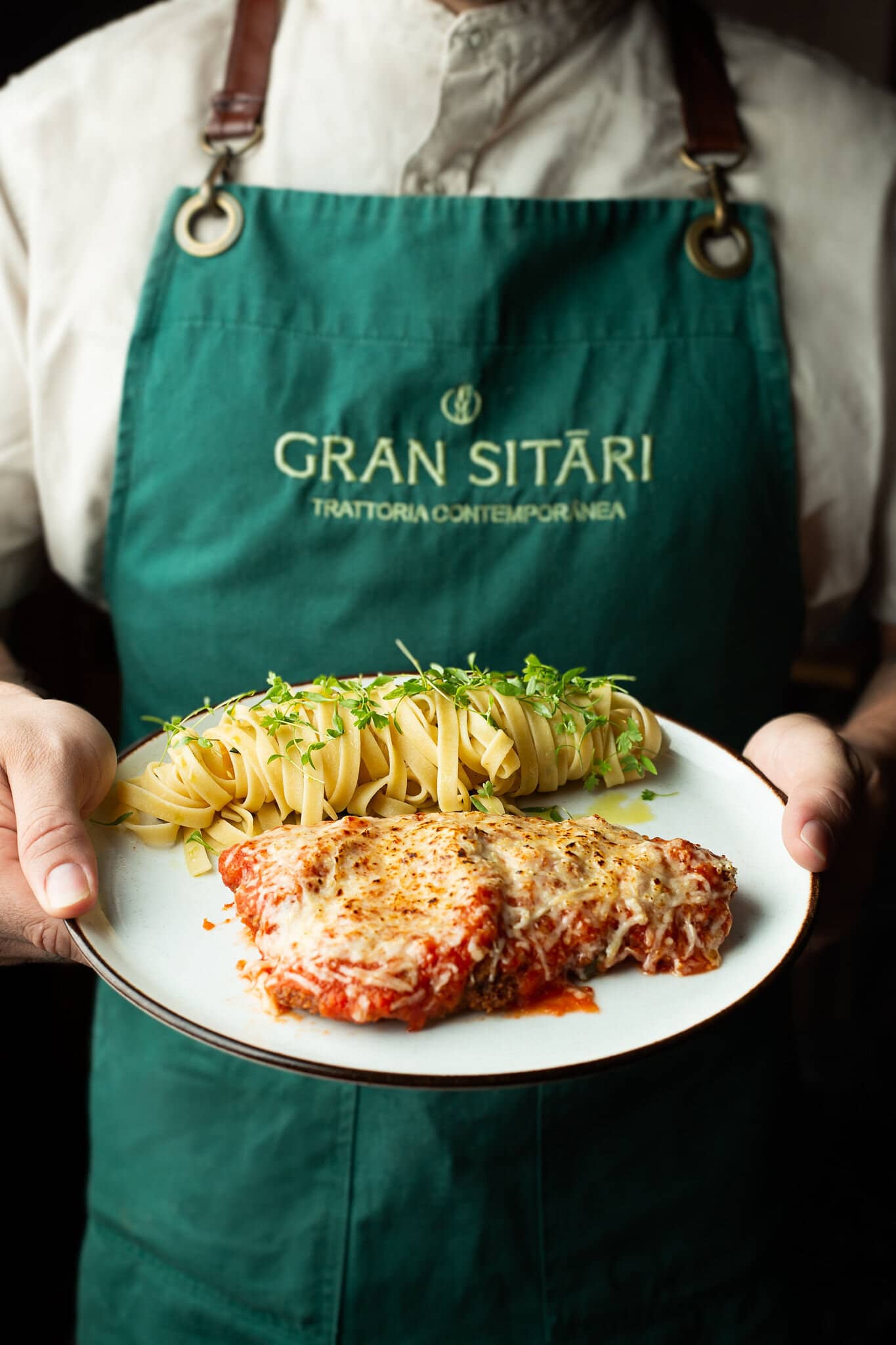 Cook in a green Gran Sitári apron holding a plate of parmigiana with tagliatelle.