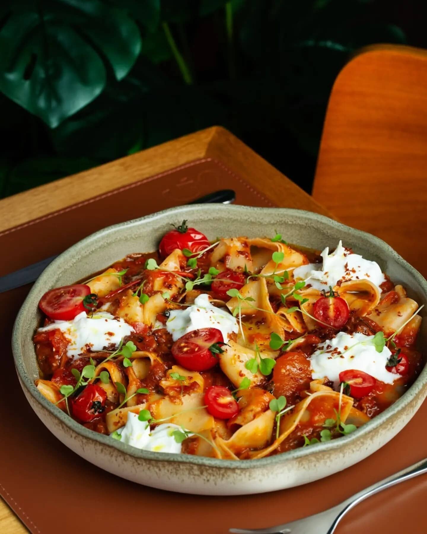 Pappardelle com ragù, burrata e tomates-cereja em prato fundo, com folhagens ao fundo.